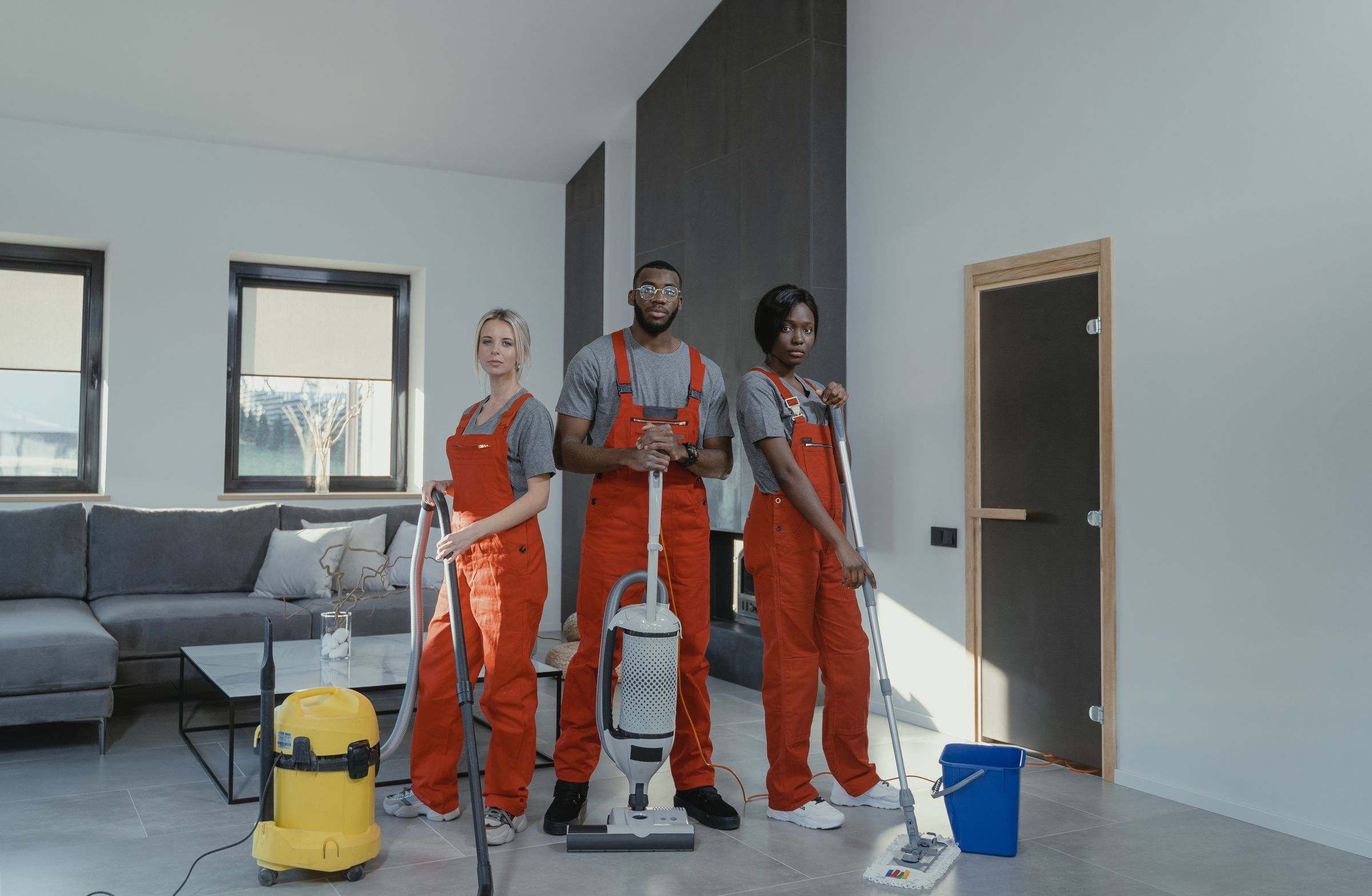 Three cleaners in red uniforms standing with a vacuum, mop and equipment in a bright modern living room