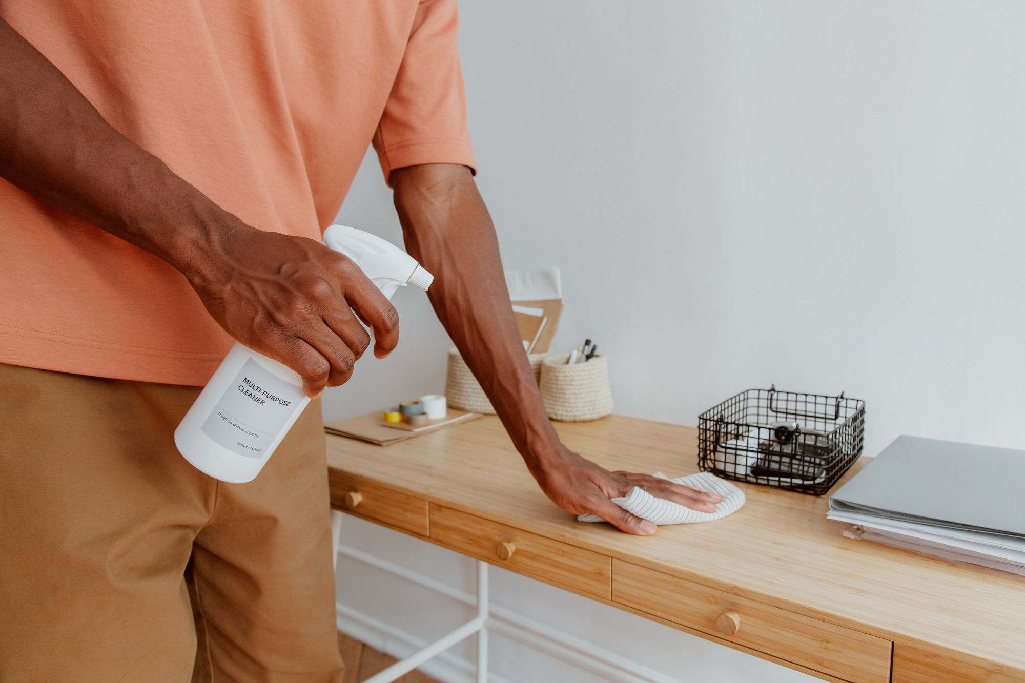 A person wiping a wooden office desk with a cloth and a bottle of multi-purpose cleaner