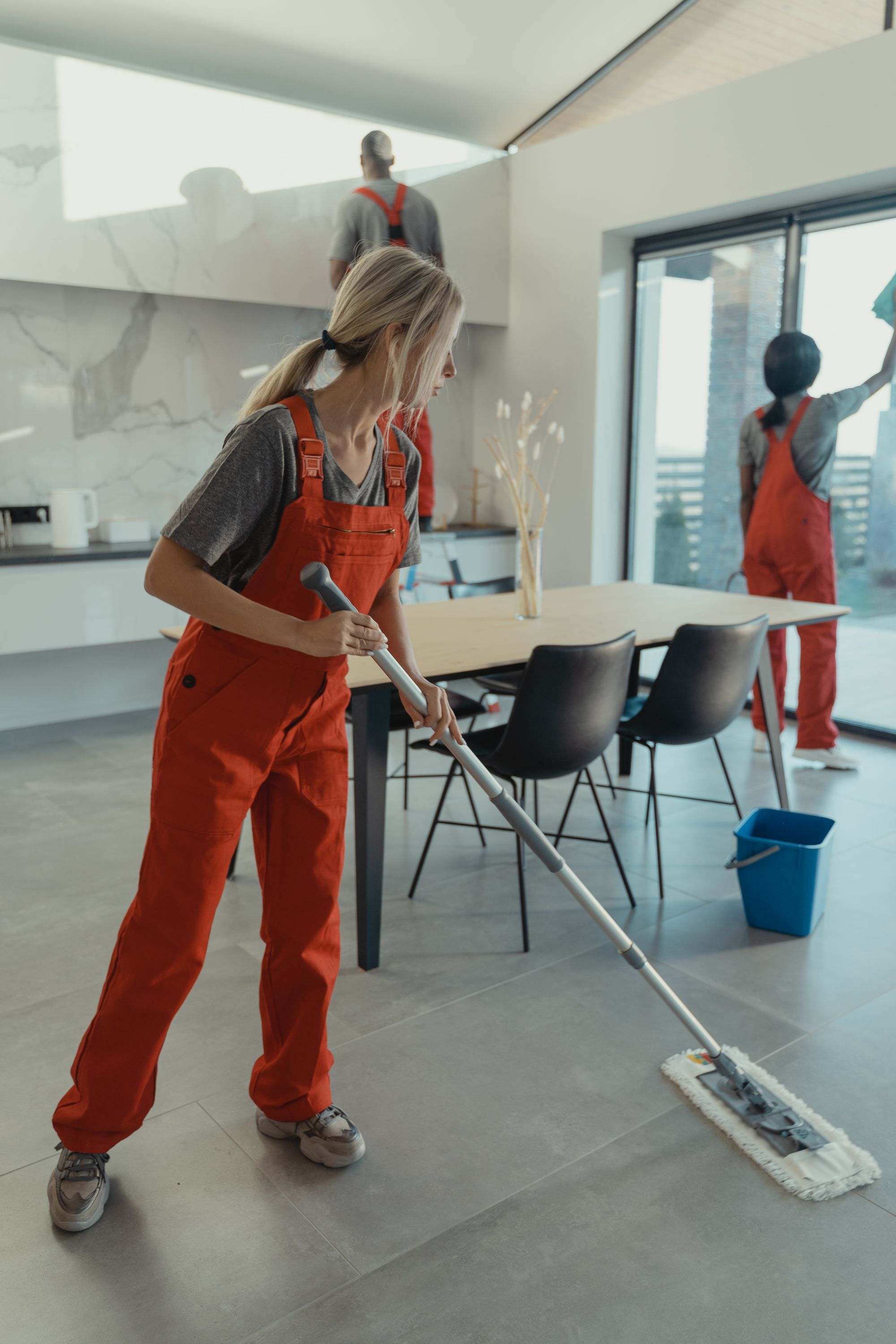A cleaner in a red uniform mopping the floor of a modern open-plan home while a colleague cleans the window